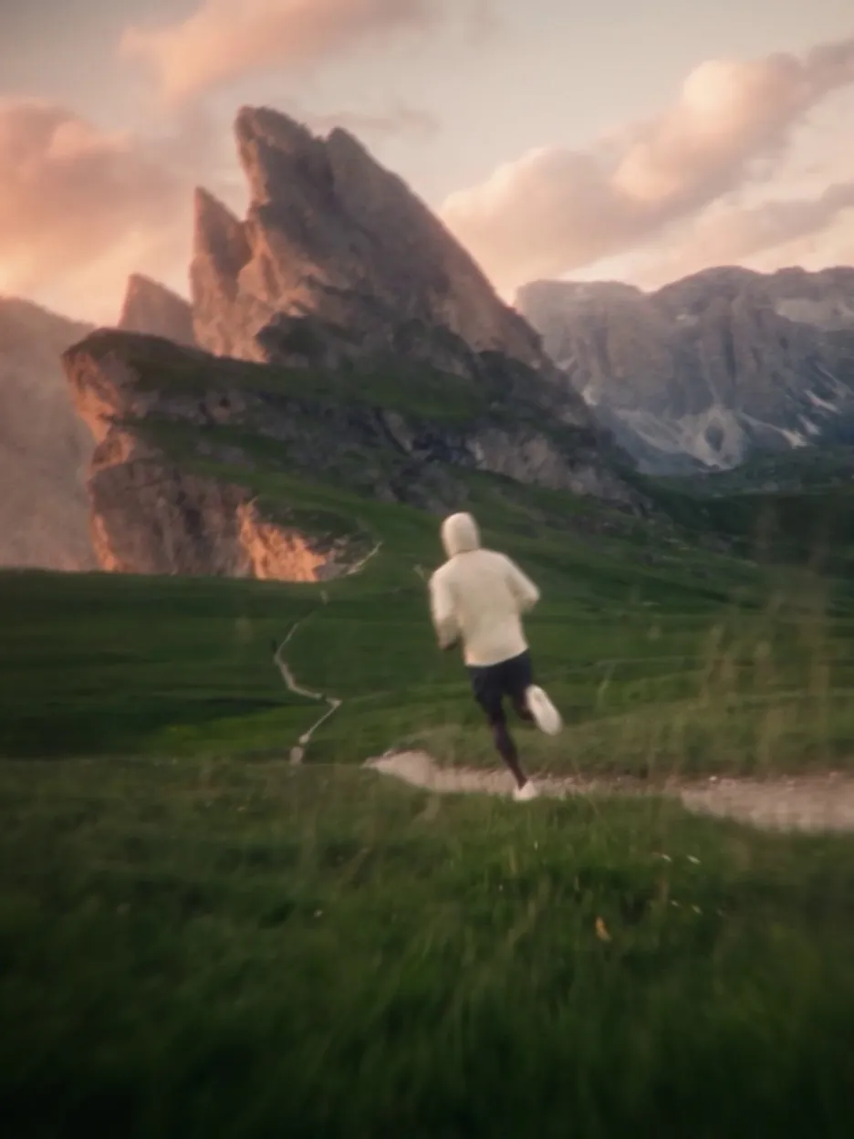 Runner in a white hoodie on a trail through the mountain pastures of the Dolomites, heading for a ridge lit by the sunset